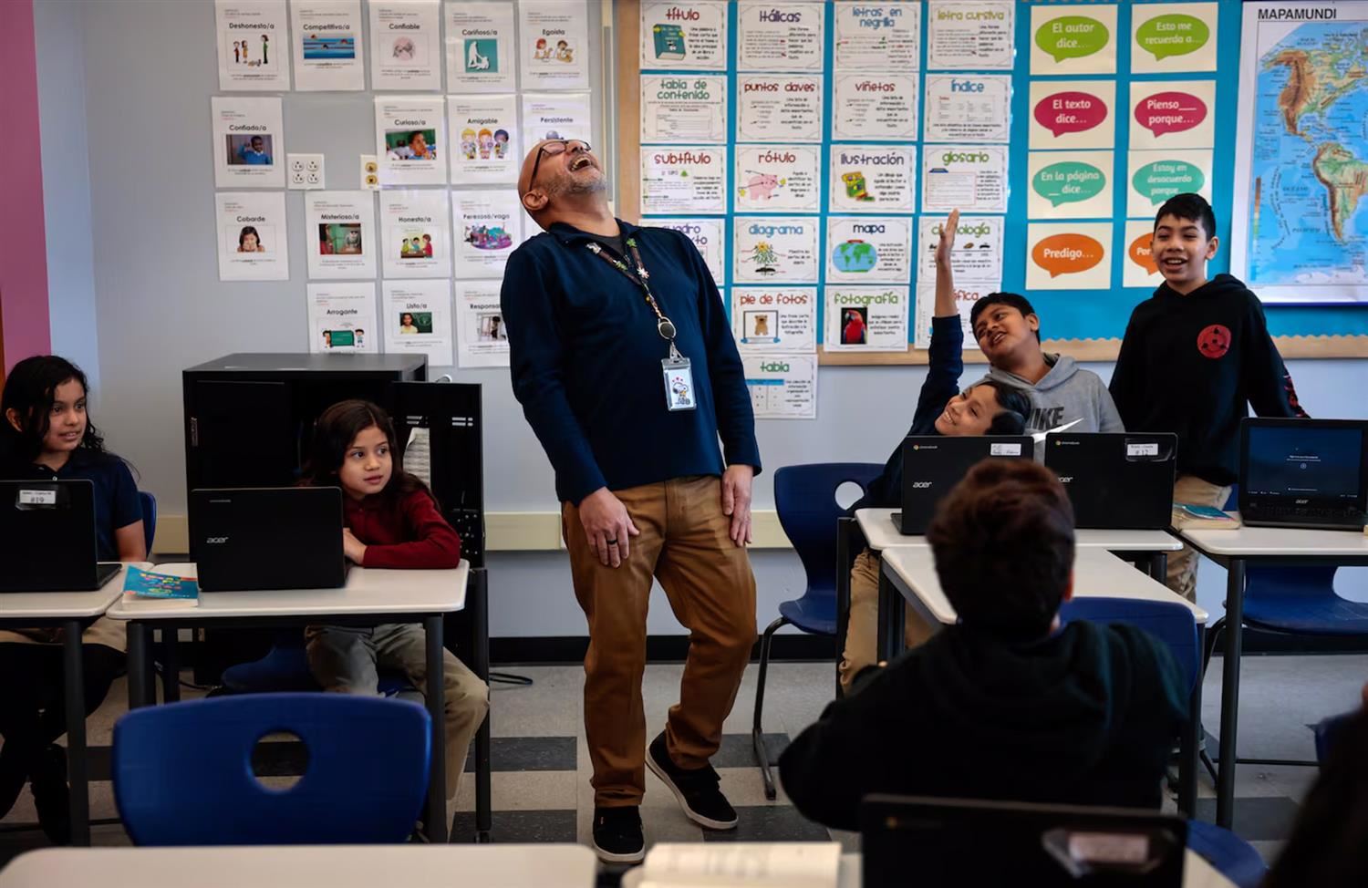 A Kelly School educator standing and smiling while teaching in a classroom. He is surrounded by elementary school students.